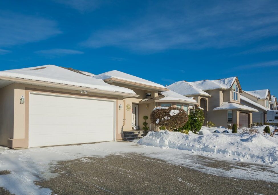 Wide garage of luxury house with driveway and front yard in snow. Street of residential houses on winter sunny day